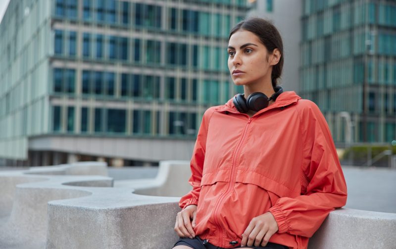 Young female in windbreaker leans with elbows on stone does exercises outdoors motivates to lead healthy lifestyle goes in for sport on city street looks forwards poses against urban building behind
